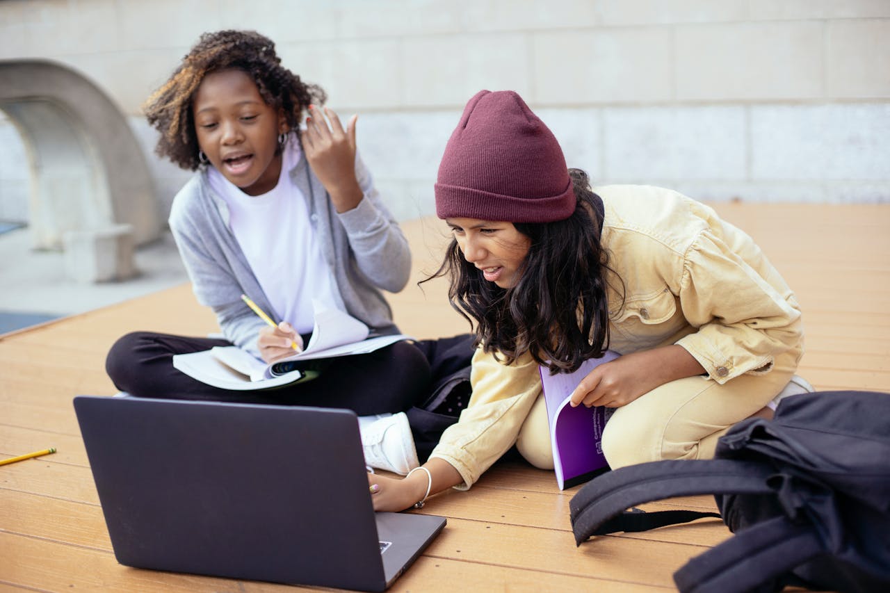 Attentive diverse schoolchildren watching portable computer while doing homework and talking on wooden platform in daylight